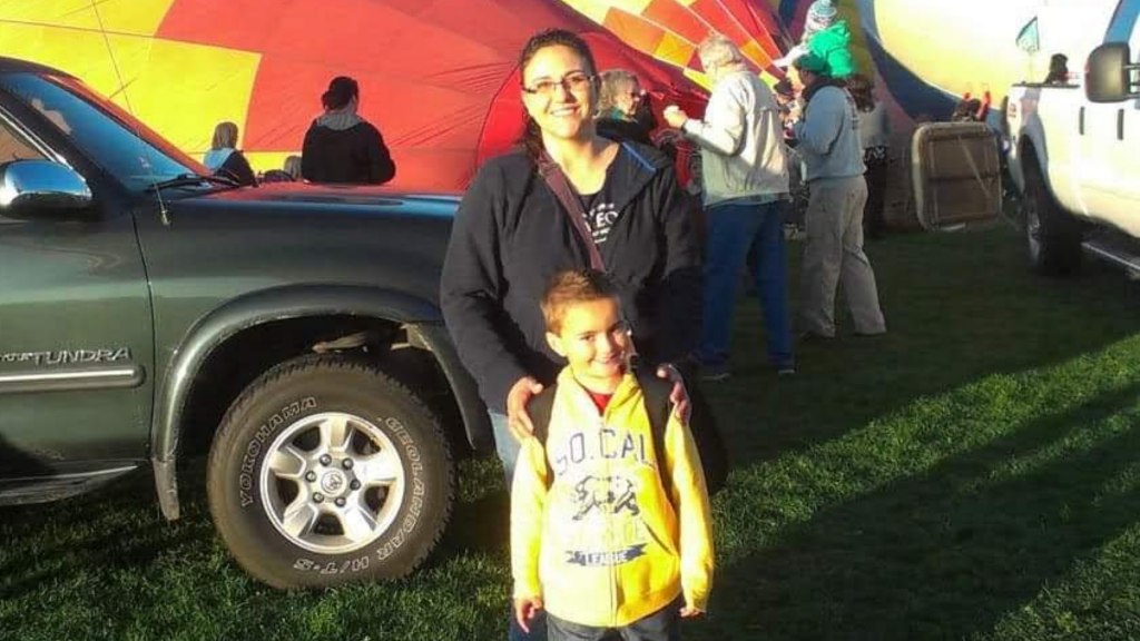 A photo of Timothy Montoya and his mother Elizabeth at a balloon rally. Timothy is smiling, standing in front of his mother as hot air balloons inflate on the ground behind them.